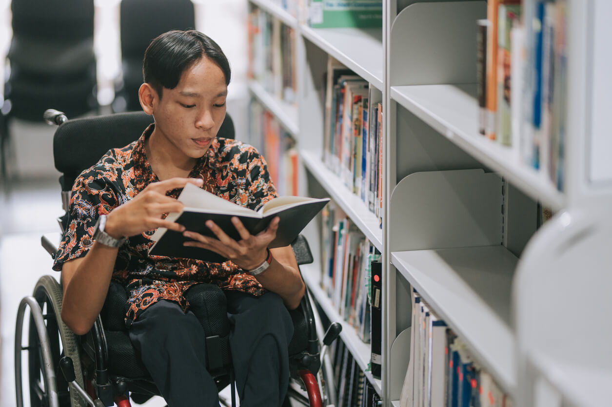 Asian teenage boy with disability in wheelchair reading book in library