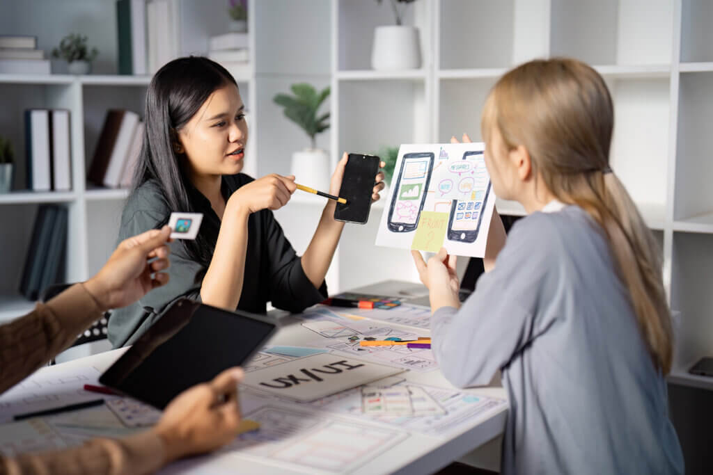 An Asian female UX designer discusses mobile app prototypes, as a white female dev listens.